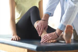 A woman is sitting on an examination table in a doctor's office. She has her shoes and socks off and the doctor is examining one of her ankles.