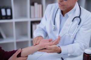A male orthopedic doctor in a white lab coat and stethoscope examining the wrist and arm of a patient for hand arthritis.
