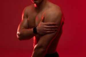 A shirtless man with a muscular build holds his arm, showing discomfort from a muscle strain.