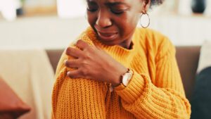 A woman sits on a couch at home, holding her shoulder with both hands as she experiences pain and discomfort.
